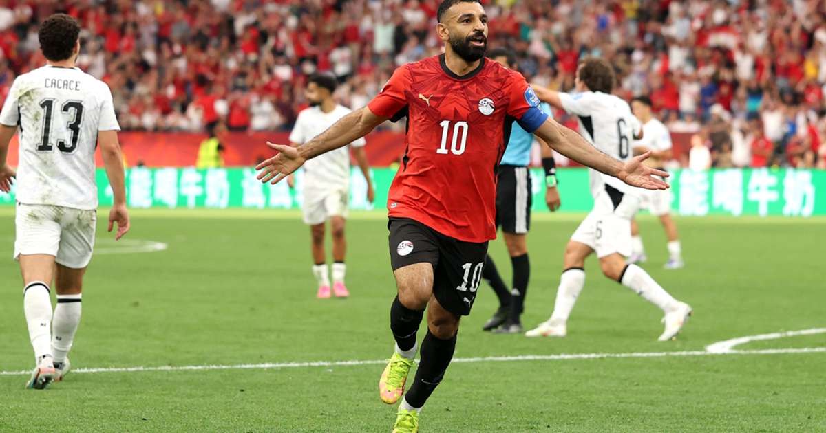 Un joueur en maillot rouge célèbre un but tandis que des joueurs en maillots blancs réagissent en arrière-plan