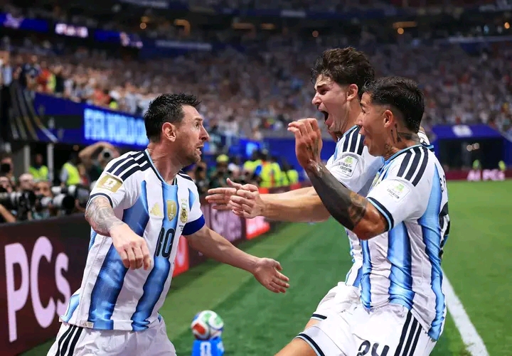 Trois joueurs de football en maillot bleu et blanc célèbrent un but sur un terrain de stade rempli de spectateurs