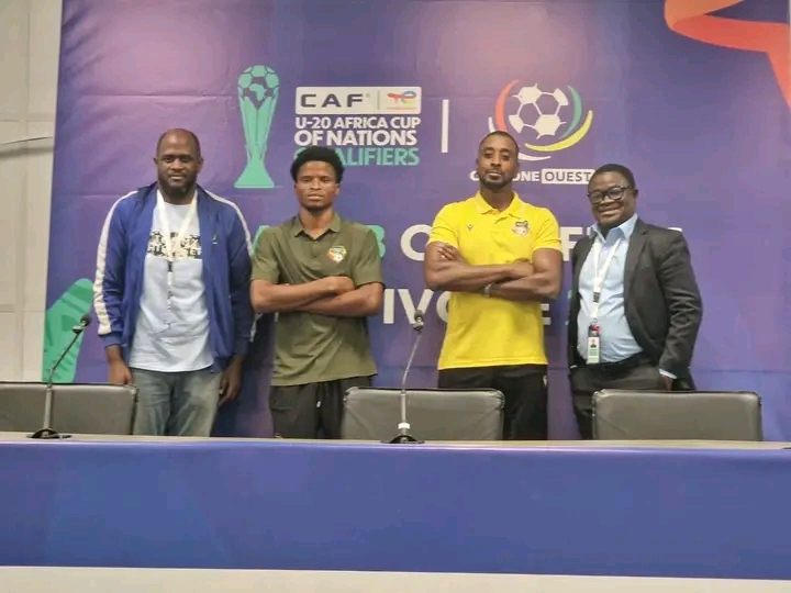 Quatre hommes debout devant une table avec microphones sur fond bleu affichant des logos de football et de compétition