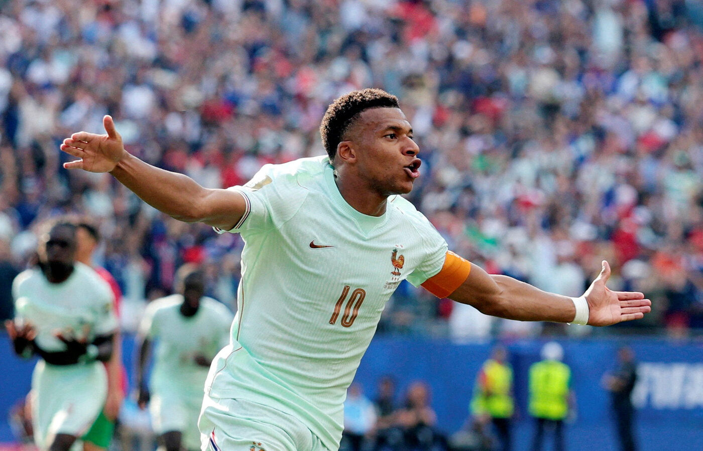 Un joueur de football en maillot blanc célèbre un but avec les bras ouverts devant une foule en tribunes.