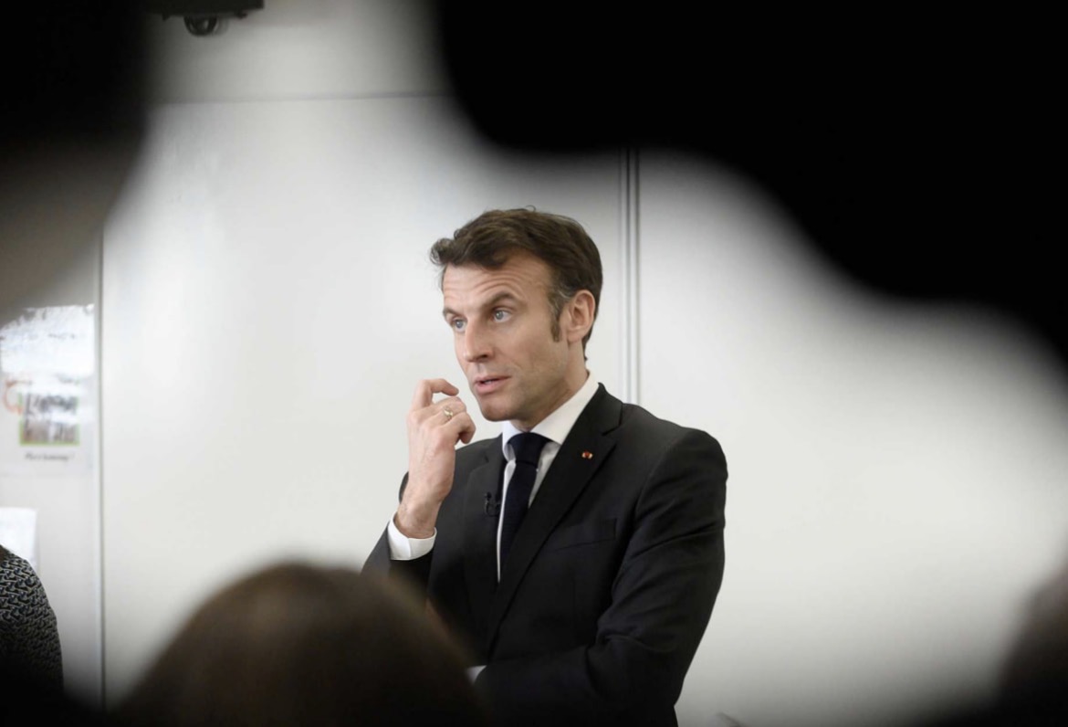 Un homme en costume noir parle devant un groupe, vu à travers une silhouette floue au premier plan