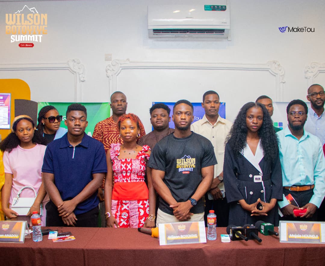 Dix jeunes adultes posent debout derrière une table lors d’un événement avec logo « Wilson Botoyiye Summit » visible au mur
